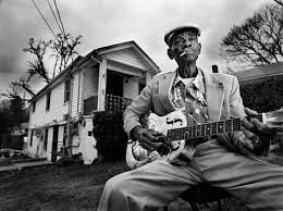 older man holding guitar in front of a small white house