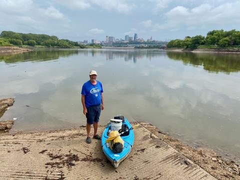 Johnny Lynn standing next to his teal colored kayak with Kansas City in the background