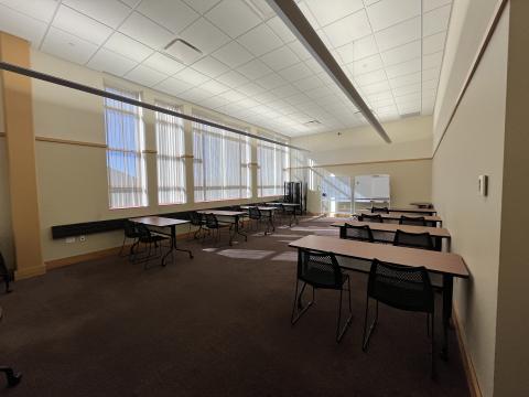 photo taken from the entrance of the room. rectangular room with windows along one side. 8 tables set up classroom style with 2 chairs at each. White boards on the far end.