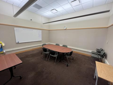 photo taken from the door way of the room, tables are arranged in the center of the room with 6 chairs around them, extra table and chairs pushed against the wall