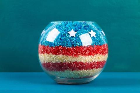 photo of a clear round vase with layers of red, white, and blue rice in it