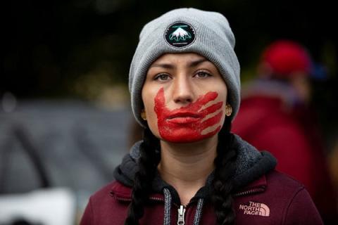 native woman with red handprint across her face, covering her mouth, wearing a grey stocking cap, staring defiantly at the camera