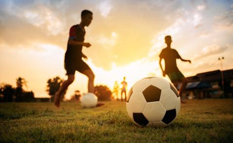 two boys kick a soccer ball in a grassy field in front of a sunset