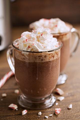 two clear glasses with handles sit on a wooden table. they are filled with hot chocolate, and topped with whipped cream and sprinkles of peppermint.