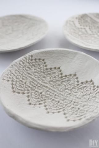three white bowls made with air-dry clay sit on a table. The bowl closest to the viewer has a stripe of a complex and intricate lace pattern stamped into the center. 