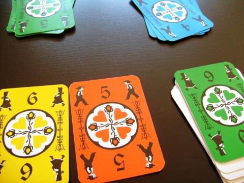 a set of Dutch Blitz cards lays scattered on a table. the viewer's hand has an orange 5, a yellow 6, and a green 5 in front of them ready to play. you see a stack of green cards a stack of blue cards further away on the table.