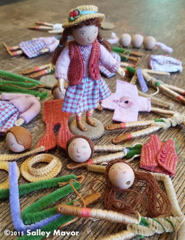 a small figurine stands among other doll parts. she is wearing a light purple sweater, blue and red checkered skirt, and a red vest, all handmade out of felt.
