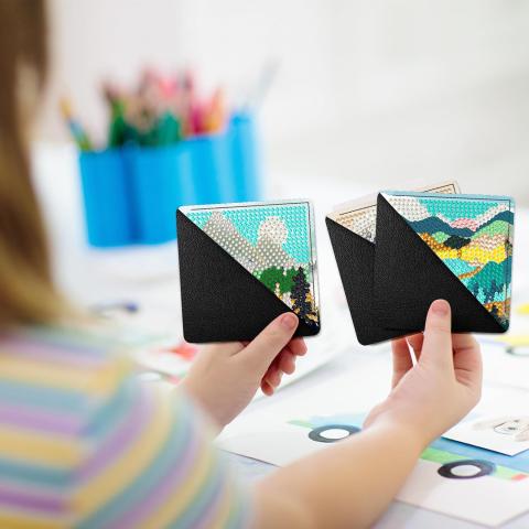 a young woman holds three corner bookmarks each depicting a different mountain scene made up of blue, white, yellow, and black diamond dots