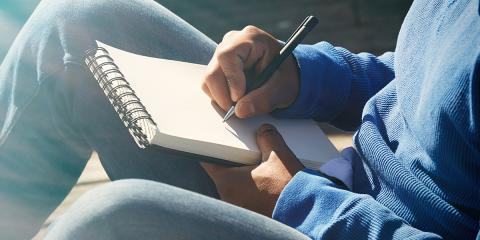 a young person wearing a blue hooded sweatshirt and jeans is sitting down in front of a light source, maybe a window, holding a spiral notebook and a pen. You cannot see their face, just their body and hand as they begin to write