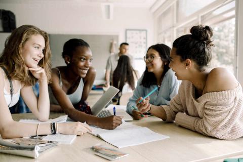 a group of young women sit around a table, smiling, as if sharing ideas.