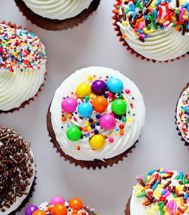 A chocolate cupcake with vanilla frosting. It is decorated with rainbow sprinkles and multicolored candies.