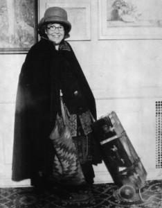 black and white photo of Anita Burnham wearing a hat and a cape, holding the handle of a piece of 1930s luggage